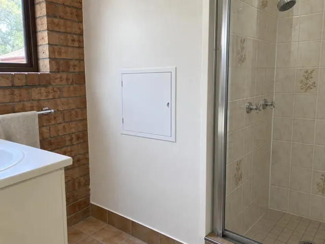 White access panel in a painted bathroom wall beside a tiled shower and brick feature wall in Kellyville